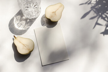 Summer stationery still life scene. Glass of water and cut pear fruit in sunlight. White table. Blank paper card, invitation mockup scene, olive branches shadows. Flat lay, top view, no people.
