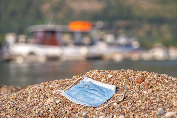 A discarded medical face mask contaminates a beautiful sandy pebble beach, with fishing boats in the background. Budva, Montenegro