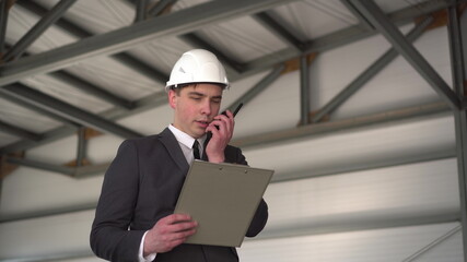 A young man in a helmet with documents speaks on a walkie-talkie at a construction site. The boss in a suit keeps records of documents on architecture. A man looks around.