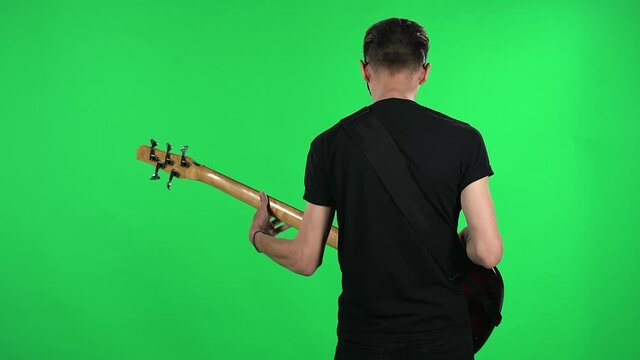 Portrait professional musician playing the electric guitar. Young guy with glasses and a black T shirt on a green screen in the studio. Back view.