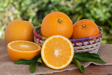 Ripe whole oranges and cut pieces in basket on wooden table with blurred green background. Copy space.