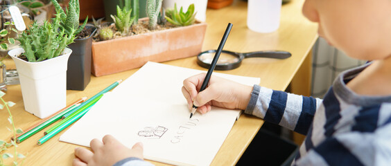 Hands of young child drawing and writing word 'cactus' on paper notebook on wooden table with color...