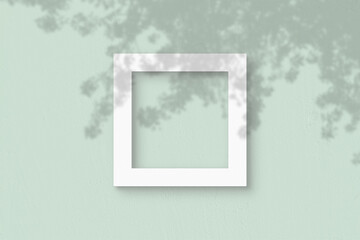 The square sheet of white textured paper on the light green wall background. Mockup with an overlay of plant shadows. Natural light casts shadows from the leaves of an exotic plant. Flat lay, top view