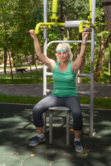 Slender elderly woman with gray hair conducts individual fitness classes on a simulator in a city park