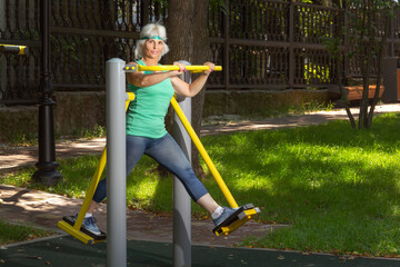 Fototapeta premium Slender elderly woman with gray hair conducts individual fitness classes on a simulator in a city park. Copy space