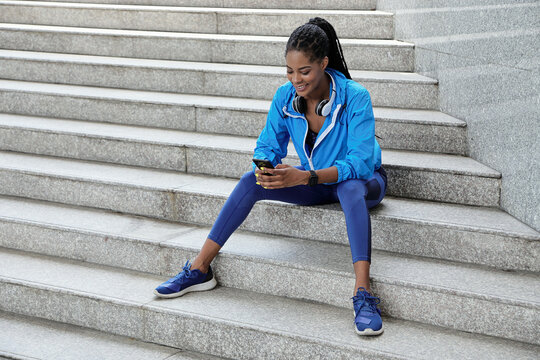 Happy pretty young African-American woman sitting on steps outdoors and choosing training program via smartphone application