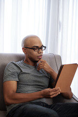 Handsome young Black man in glasses sitting on sofa and reading interesting book on tablet computer