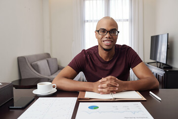 Potrait of handsome smiling young Black businessman in glasses sitting at desk at home and working with financial documents