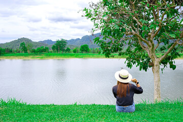 Woman wear a hat sitting by the big lake with trees and mountains view. Vacation time concept.