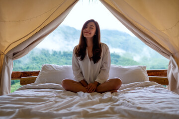 Fototapeta na wymiar Portrait image of a young woman sitting on a white bed in the morning with a beautiful nature view outside the tent