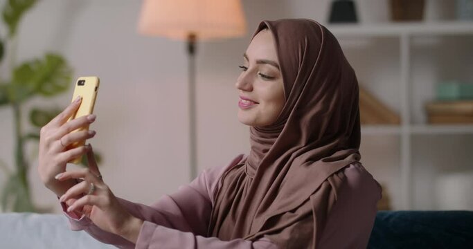 Young attractive muslim girl sitting on couch, looking at mobile phone camera and taking selfies for social media. Muslim woman having active social life 4k footage
