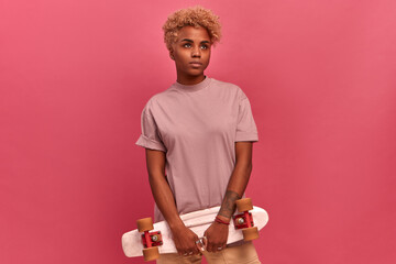 Pensive African American teenager with a skateboard on a pink background. Girl with dark skin in casual clothes stands and thinks who will teach her to ride penny board. Concept of learning new skills © antianti