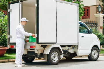 Delivery man in white uniform taking plastic crate with glass milk bottles out of van trunk