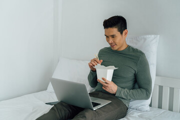 Portrait of Asian man eating takeout food watching laptop on bed. New lifestyle, food order from modern technology concept.