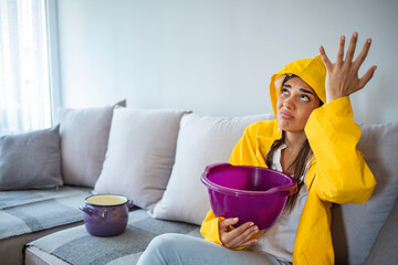 Worried Woman in Raincoat Holding Bucket While Water Droplets Leak From Ceiling in Living Room....