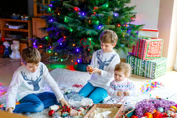 Fototapeta na wymiar Two little kid boys and adorable baby girl decorating Christmas tree with old vintage toys and balls. Family preaparation celebration of family holiday. Three children, brothers and sister at home.