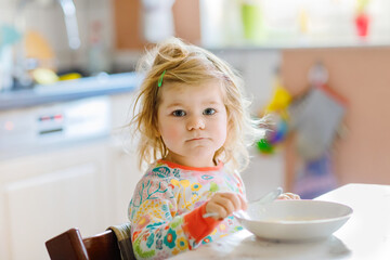 Adorable toddler girl eating healthy porrige from spoon for breakfast. Cute happy baby child in colorful pajamas sitting in kitchen and learning using spoon.
