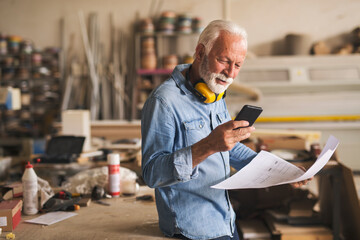 Likeable older carpenter is looking at the cellphone and smiling