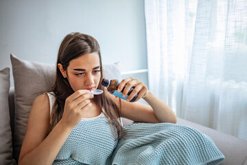 Exhausted young woman preparing to drink spoonful of cough syrup while suffering from flu at home....
