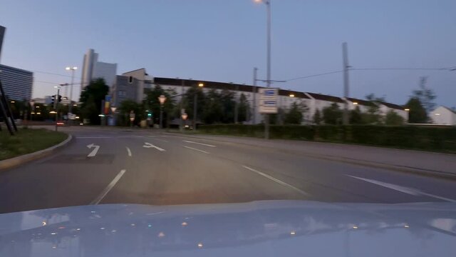 Drive through Munich, a bavarian capital trip by car at the evening. Speeded up from the engine hood view at the late evening with illuminated lanterns and modern buildings.