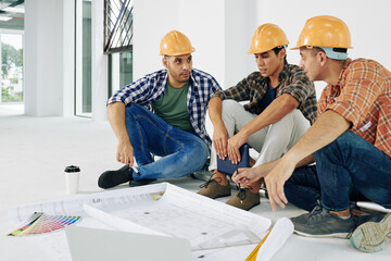 Young serious builders in hardhats sitting on the floor and discussing how to renovate apartment