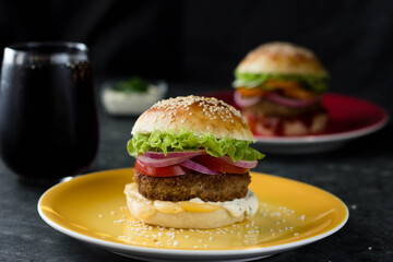 A home-made hamburger is on a bright plate, another is behind it, and a glass of coke is next to it