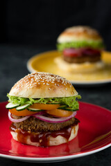 A home-made hamburger is on a bright plate, another is behind it, and a glass of coke is next to it