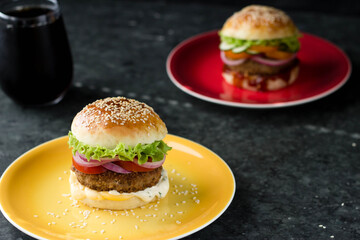 A home-made hamburger is on a bright plate, another is behind it, and a glass of coke is next to it