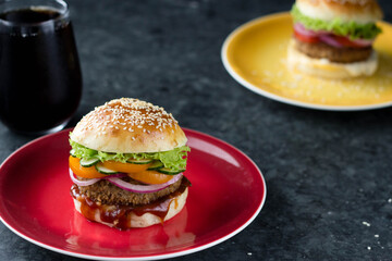 A home-made hamburger is on a bright plate, another is behind it, and a glass of coke is next to it