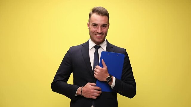 happy elegant businessman in suit holding clipboard smiling and holding hands in pocket, standing on yellow background