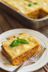 Homemade lasagna ready to eat in metal baking tray served with basil leaves, tomatoes, and turquoise tablecloth on brown wooden background. One portion on the white plate with fork. Top view.