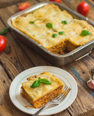 Homemade lasagna ready to eat in metal baking tray served with basil leaves, tomatoes, and turquoise tablecloth on brown wooden background. One portion on the white plate with fork. Top view.