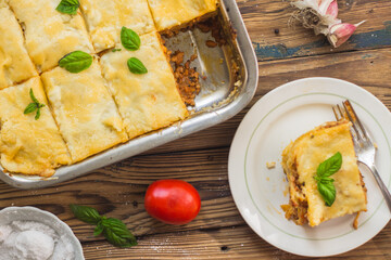 Homemade lasagna ready to eat in metal baking tray served with basil leaves, tomatoes, and turquoise tablecloth on brown wooden background. One portion on the white plate with fork. Top view.