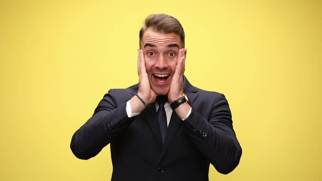 excited young businessman in suit hearing news, holding hands in the air, holding hands on face, smiling and pointing fingers on yellow background