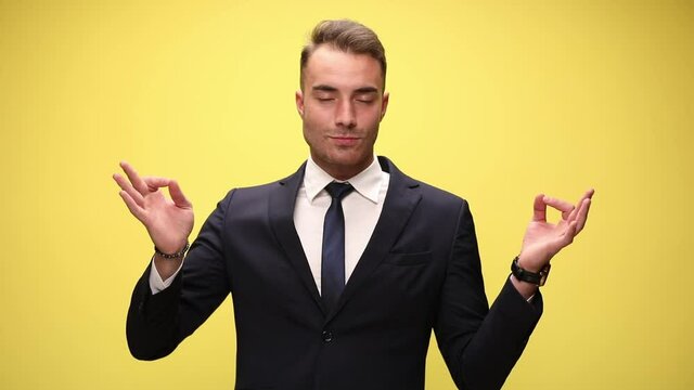 radiant young businessman in suit, smiling, holding fingers in a gian mudra position, relaxing arms and body and meditating on yellow background