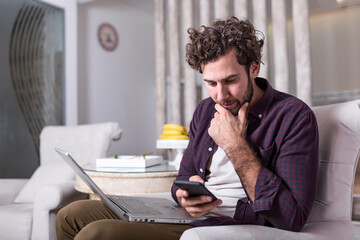 Young handsome man making video call with friends while sitting on sofa at his modern home.Concept of happy business people. Business man at home checking statistics on mobile phone and laptop