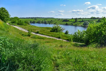 Dirt road among green hills on the shore of a pond with blue water. Bright natural panorama with a lake in the middle of juicy young grass on a spring-summer day