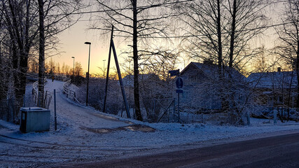 Winter sunset landscape with snow, trees, wooden houses and a pedestrian bridge.