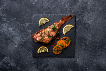 Baked sea bass with tomato pepper lemon and herbs on black slate stand on dark concrete background. Perch fish cooked in the oven. Copy space Top view