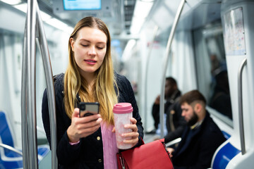 Portrait of positive girl wearing warm coat traveling in metro car and using mobile phone