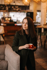 Close up of face of young woman holding cup of tasty hot coffee in hand while standing in restaurant. Caucasian lady posing with glass of beverage in cafe with modern dark interior.