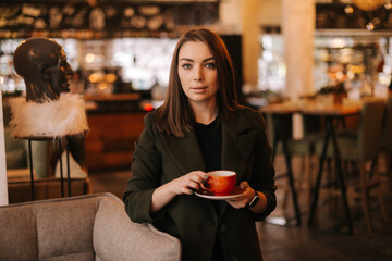Fototapeta premium Beautiful young woman holding cup of tasty hot coffee in hand while standing in restaurant. Caucasian lady posing with glass of beverage in cafe with modern dark interior.