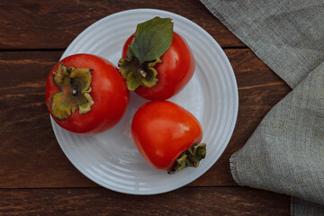 Ripe, red, juicy persimmon with a green leaf on a white plate with a linen napkin on a brown wooden background top view, the concept of choosing healthy seasonal fruits