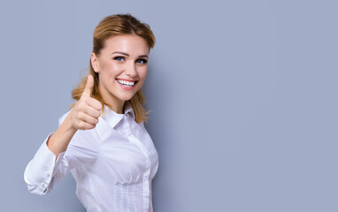 Portrait of happy smiling young excited businesswoman, showing thumb finger up or like hand sign gesture. Success in business concept studio shot. Isolated over grey color background. Copy space area.