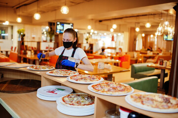 Waiter in protective mask cutting pizza in pizzeria.