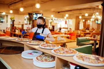 Zelfklevend Fotobehang Waiter in protective mask cutting pizza in pizzeria. © AS Photo Family