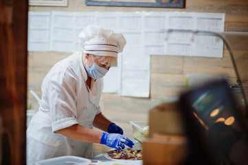 Pizza maker in protective mask working in the pizzeria.