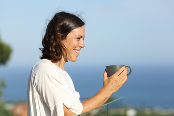 Happy adult woman with coffee looking away in a balcony
