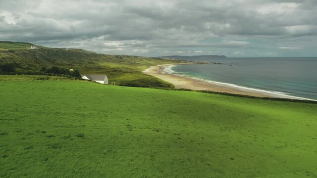 Beach lodge aerial shot: green grass meadow near sea bay and lonely White Rocks. Grey clouds hover over ocean water, sandy shore. Tranquil landscape of nature in Northern Ireland. Footage in 4k, UHD