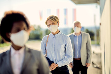 Businesswoman with face mask standing at safe distance while waiting in a line outdoors.